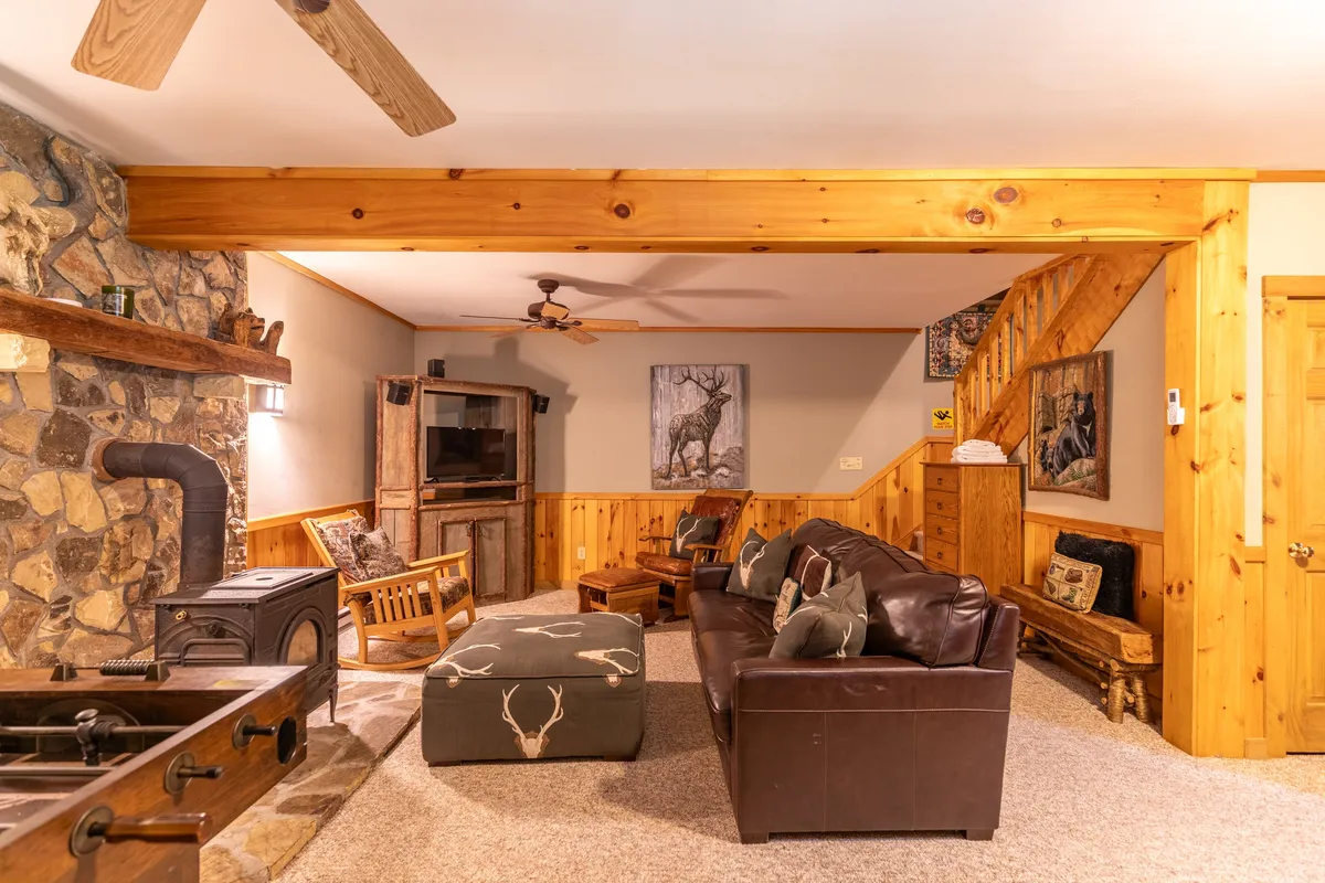 A rustic living area with a prominent stone fireplace, leather sofa, and wooden beams. The space is accented with decorative wall art and a staircase, giving a welcoming, lived-in feel ideal for gathering and unwinding. The arrangement suggests possibilities for socializing, reading, or enjoying a casual game night.
