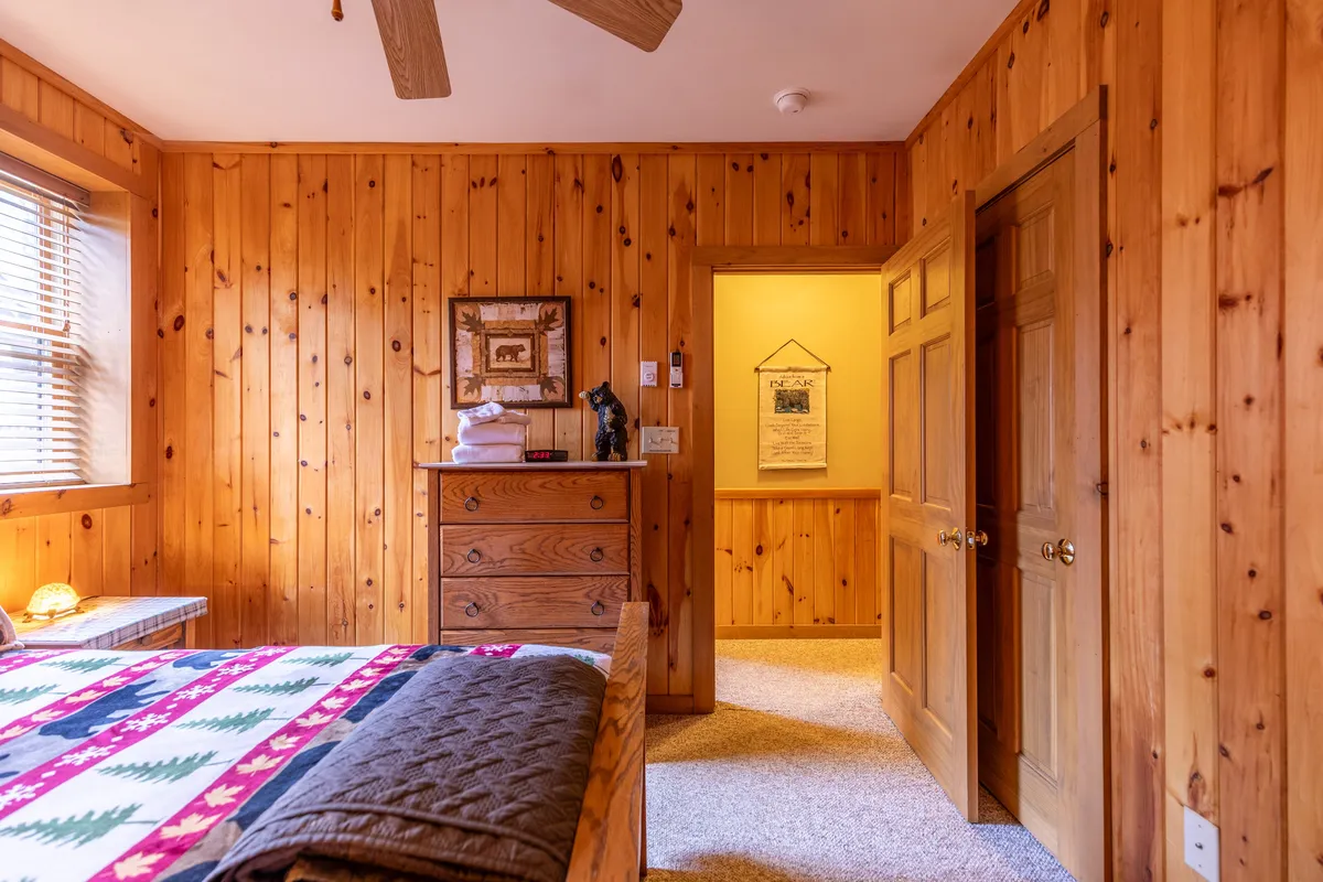 A bedroom featuring a patterned quilt on the bed, wooden dresser, and framed artwork. Through an open door, soft natural light filters in, suggesting a peaceful connection to the outdoors and mornings spent waking up to nature.