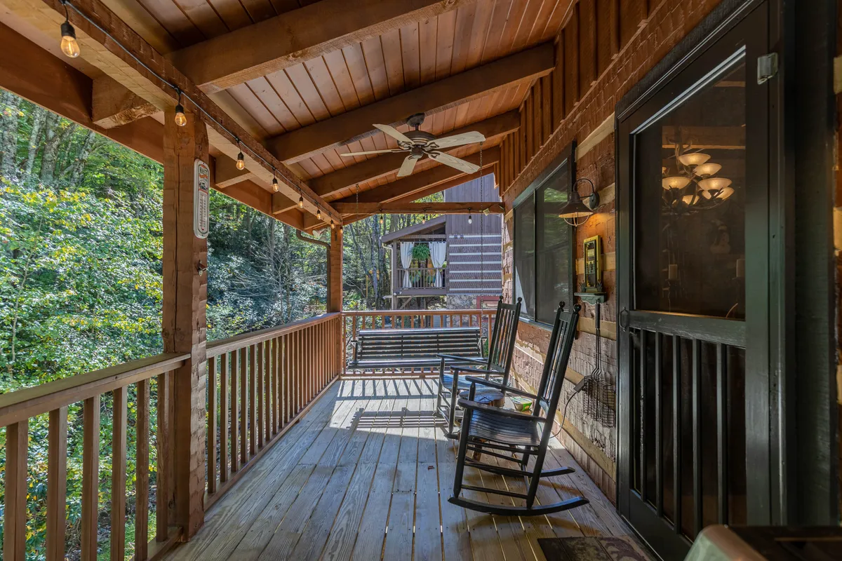 This inviting exterior shot shows a spacious covered porch with classic wooden rocking chairs, ceiling fan, string lights, and a sturdy wooden railing. The porch overlooks lush greenery, creating a relaxing atmosphere ideal for unwinding after a day of adventuring with friends.