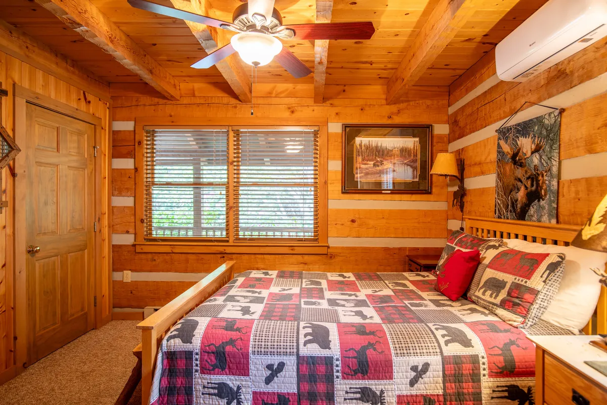 A cozy bedroom with a queen-sized bed dressed in a patterned quilt, set against warm wooden walls. Sunlight filters through windows with blinds, creating an inviting, restful atmosphere. The room is uncluttered, emphasizing comfort and privacy.