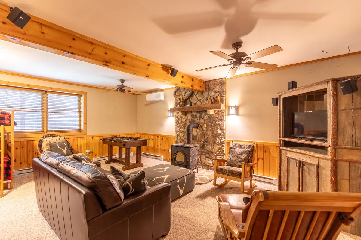 A spacious rec room with a stone fireplace, wooden beams overhead, plush seating, and an entertainment center. The setup includes a coffee table, inviting a group to gather for board games or movie nights. Natural light filters through windows, giving the area a warm, social ambiance.