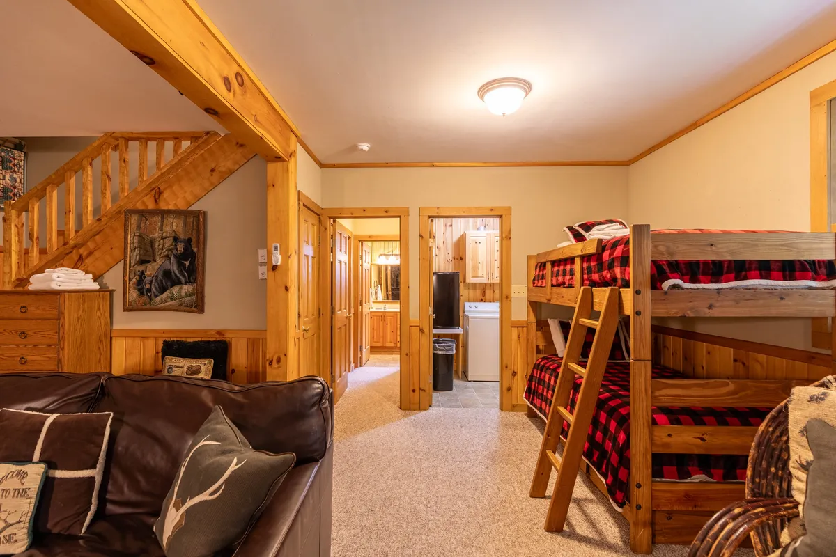 A cozy lower-level living space featuring bunk beds against the wall, a comfortable sofa, a staircase leading upstairs, and rustic wooden furniture. Framed artwork adds a playful touch, and the open layout makes it perfect for gatherings and games.