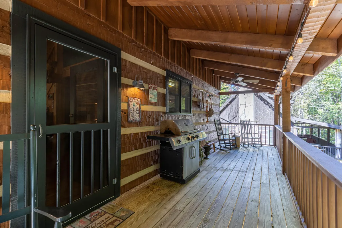 A cozy exterior porch scene featuring a grill, a rocking chair, wooden railing, and decorative wall signs. The space is inviting for both relaxing and socializing, with lush green surroundings visible in the background. This image also subtly hints at the covered nature of the deck and the casual, welcoming mountain retreat atmosphere.