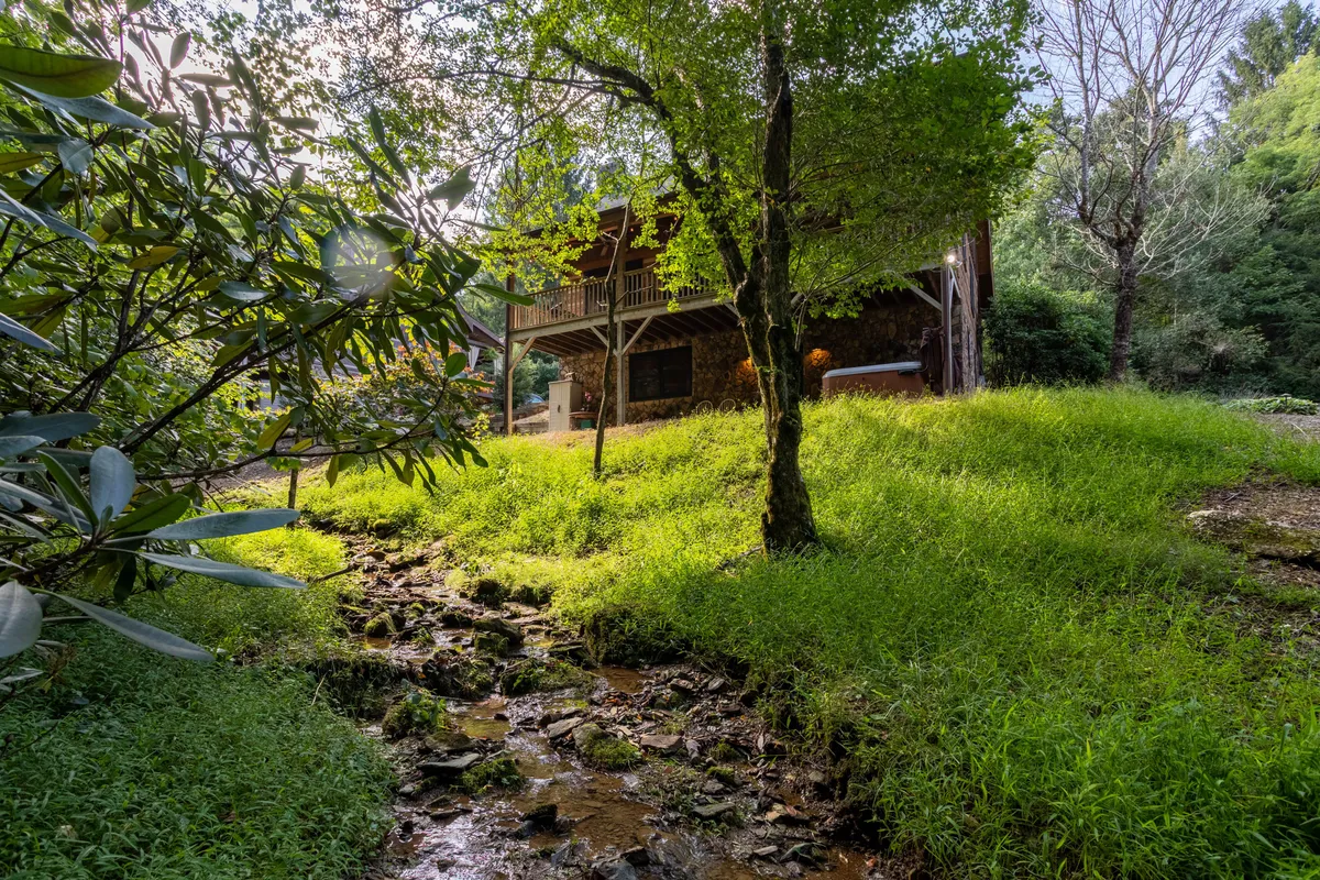 A picturesque view of the house on stilts, with a gentle, flowing creek in the foreground and lush grass and trees all around. The scene is serene and naturally beautiful, emphasizing the seasonal creek and wooded setting that enhance the cabin’s outdoor appeal.