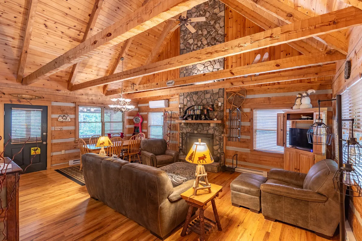 A warm, inviting great room featuring a striking stone fireplace as the focal point, surrounded by comfortable seating. Exposed wooden beams and log walls define the space, with a dining area and rustic wooden table visible beyond the living area.