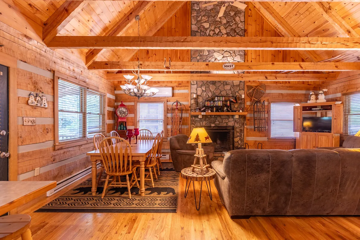 A rustic dining area with a substantial wooden dining table set for a group, positioned close to a comfortable sofa and a stone fireplace. Decorative lighting and wood accents enhance the cozy, communal ambiance.