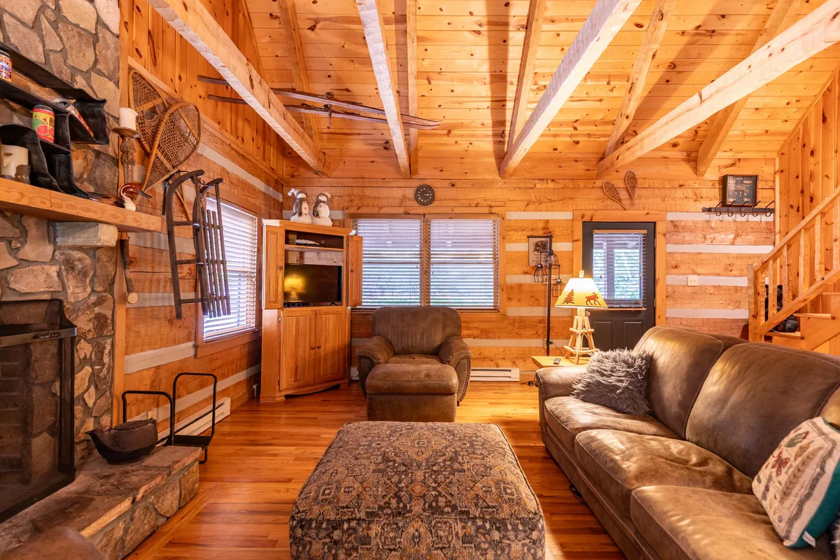 A warmly lit living area with a prominent stone fireplace, exposed wooden beams, and cozy comfortable seating. The decor features rustic accents and large windows that suggest views of the natural surroundings, while the arrangement invites relaxation and social connection.