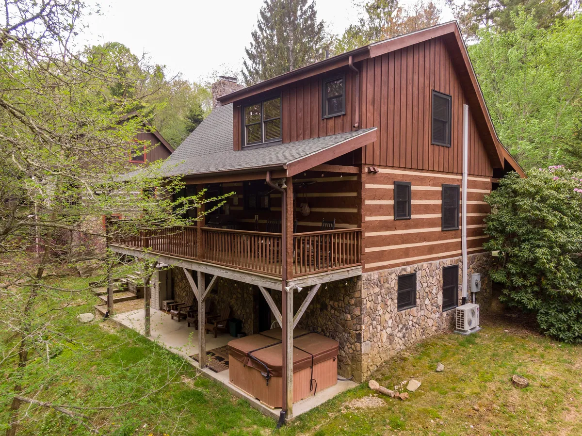 The image showcases the inviting exterior of a rustic cabin set against lush greenery, with a spacious wooden balcony, stone foundation, and a bubbling hot tub. The balcony offers sweeping views—perfect for sunset watching or stargazing—while the outdoor space invites guests to relax and connect with nature.