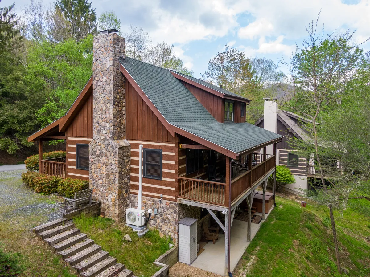 Featuring a log cabin structure with a prominent stone chimney and a wide, covered porch set amid vibrant greenery, this image highlights the inviting outdoor spaces ideal for gatherings, relaxation, and enjoying the mountain air.